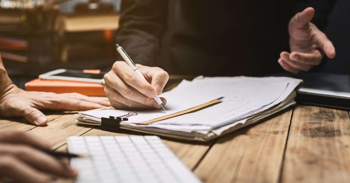 A shot of two people, focusing on their hands working at a wooden desk with paper and pencils